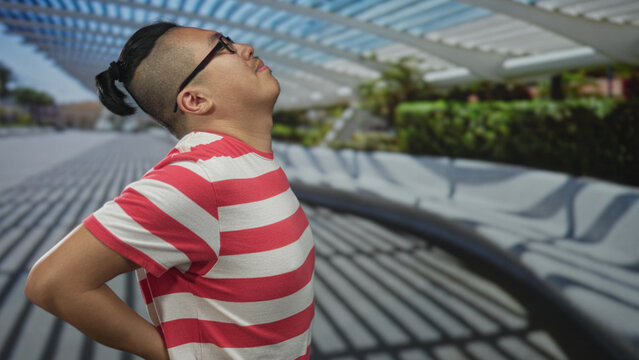 Young chinese man with glasses and manbun wearing red striped shirt leaning forward and holding lower back for backache on street under louvered canopy; discomfort.