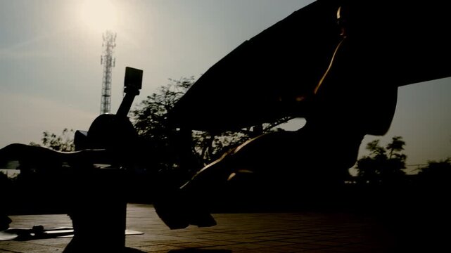 Silhouette of athletic woman training on rowing machine outdoors at sunset. Fitness, strength and endurance concept. Active female workout with dramatic backlight and copy space.