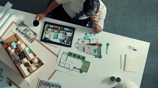Top view of engineer drink coffee and thinking about house plan at meeting table with equipment and safety helmet scatter around. Aerial view of stressful manager looking at blueprint. Alimentation.