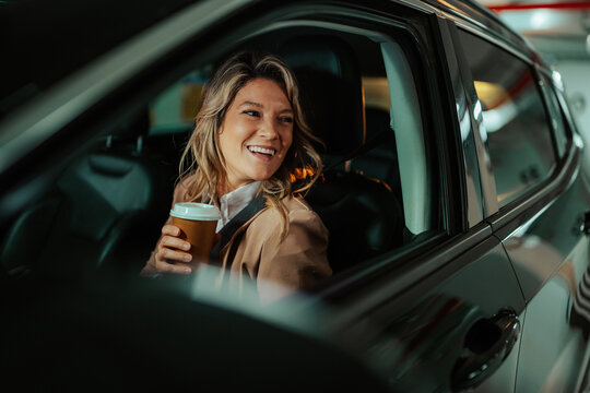 Happy woman is seated in her car after work, enjoying a take away coffee and looking completely content