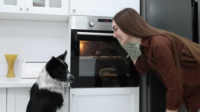 Woman taking out tray with bone shaped cookies from oven near cute dog in kitchen. Camera moving forward