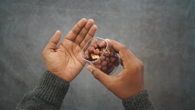 A man's hand pouring a small container of raw peanuts into his open palm with a dark background