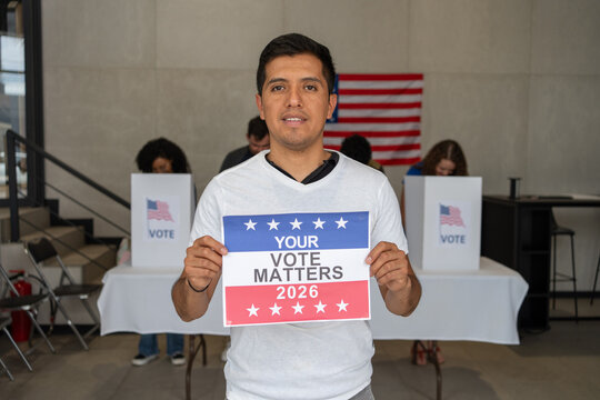 latino hispanic man holding a Your Vote Matters 2026 sign at a US election polling station. In the background, a diverse group of people votes in voting booths, democracy in America