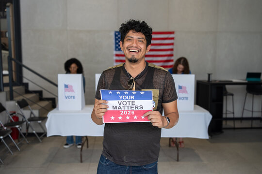 latino hispanic man holding a Your Vote Matters 2026 sign at a US election polling station. In the background, a diverse group of people votes in voting booths, democracy in America