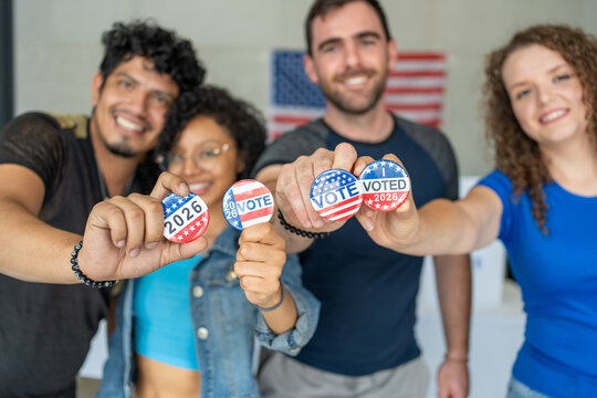 diverse group of smiling friends holding 2026 Vote election I voted buttons or pins at a US election polling station. In the background, american flag at the background, democracy in America USA