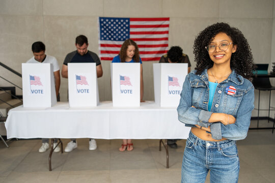 young African American woman smiles wearing a 2026 vote pin button at a US election polling station. In the background, a diverse group of people votes in voting booths, democracy in America