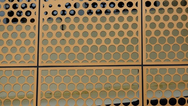 Close-up of an orange perforated metal screen with a repetitive circular pattern. A vibrant geometric architectural detail suitable for industrial backgrounds and modern design textures.