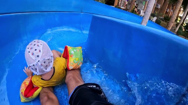POV - a kid in a panama hat and bright armbands rides along the blue water slide of the water park with an adult. Summer, family vacation, safety.
