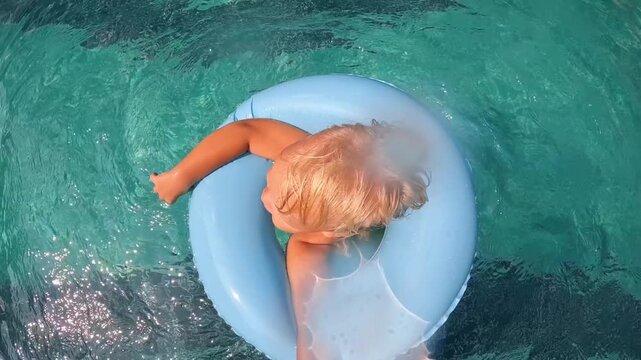 Shot from above: a child in the pool spinning in the water on a blue inflatable circle, splashes and sun glare on the surface.
