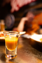 A shot glass filled with amber liquid on a wooden table in a dimly lit bar setting © chuangxin