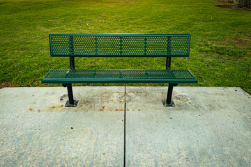 A green metal bench sits on a concrete surface facing a wide grassy area in a park the bench has a perforated pattern and is supported by sturdy black legs