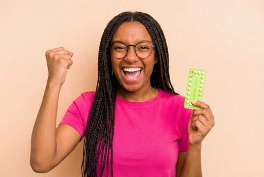 Excited african american woman holding birth control pills and raising fist in victory on beige
