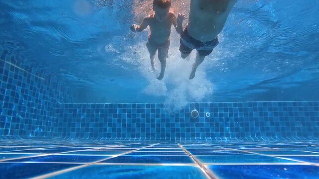 Underwater shot in the pool - father and child swimming towards the camera, surrounded by a cloud of bubbles and bright glare on the blue tiles.
