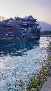 A stunning shot of the fast-flowing blue waters of the Min River in Dujiangyan, Sichuan. The background features the magnificent ancient Chinese architecture built along the riverside under a soft twi