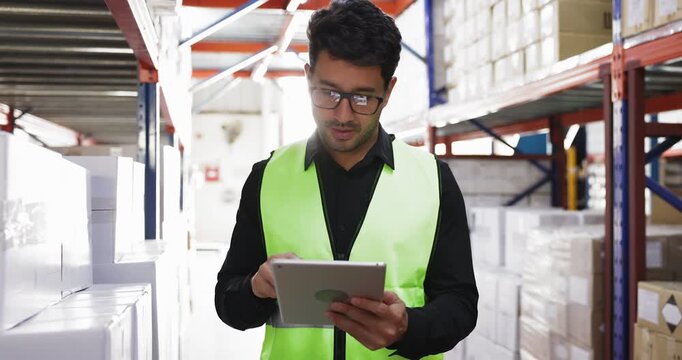 Storage worker man in glasses and yellow vest checking part numbers on boxes and tablet in warehouse, pointing at packages, counting goods, making inventory, using logistic app on gadget