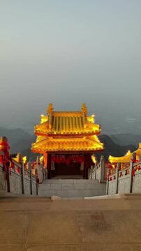 A stunning symmetrical shot of a golden-roofed Taoist temple on the summit of Mount Laojun, Luoyang. The traditional architecture is illuminated against the twilight sky, with red blessing ribbons lin