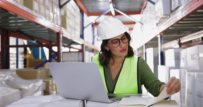 Positive young female Hispanic supervisor in glasses and hard hat working at laptop in warehouse space, using computer at metal shelves with stacked boxes, consulting notes in notebook