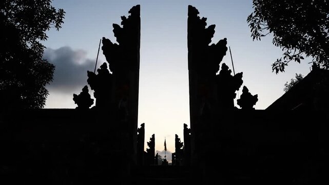 Silhouette of a traditional Balinese split gate against a bright sky at dawn, revealing a distant path.