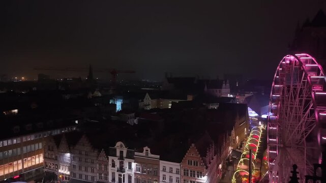 Drone pull-back revealing the Ghent cityscape with the illuminated Christmas wheel and the historic clock tower glowing in a festive winter atmosphere.