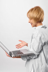 Smiling middle aged woman in white shirt holding laptop and gesturing with hand, isolated on white background. Concept of technology, communication, and remote work.
