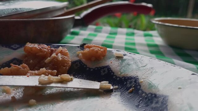 Housefly crawls over leftover food scraps on a dirty plate next to table knife, with cooking pot and checkered tablecloth in soft focus behind, illustrating hygiene and contamination, macro close shot