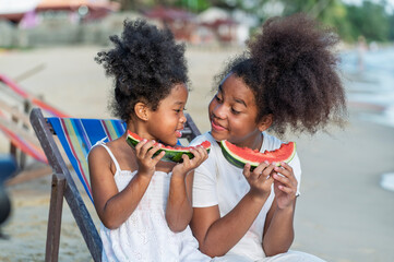 Two smiling African American girls with curly hair bite watermelon and share a happy moment of pure beauty and fun in Beach outdoors background