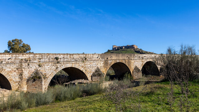 Roman arched stone bridge over the Guadiana river with medieval castle on a hill in the background in Medellin Spain ESP