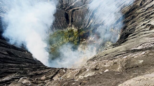 Dramatic view into the active Mount Bromo crater in East Java Indonesia, reveals thick volcanic smoke rising from steep mineral stained walls formed by powerful eruptions and ongoing volcanic activity