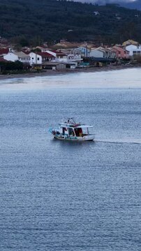 Scenic aerial view of a fishing boat moving along the shore of Acharavi village with traditional houses and hills in the background, Corfu island, Greece, Ionian Sea coastline travel destination, summ