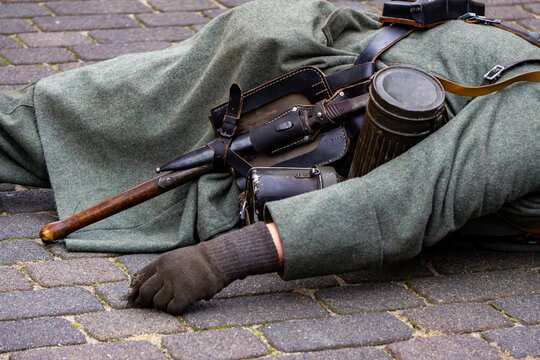 Historical reenactment, soldier in full gear lying on cobblestones, military equipment details, bayonet, gas mask canister and entrenching tool, battle staging, military history, uniform details.