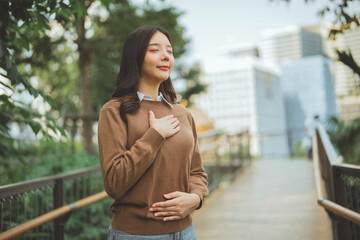 Happy Asian woman closing eyes and breathing fresh air in city park, young lady practicing mindfulness and relaxation outdoors, concept of mental health, urban wellness, stress relief, peaceful