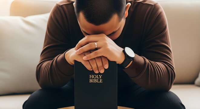 Man sitting on couch praying with hands clasped over a Holy Bible