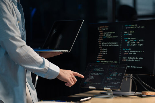 Developer collaborating across multiple screens, pointing at tablet code while holding a laptop, monitoring and refining complex software in real time

