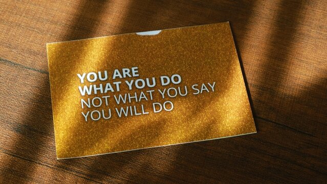 A motivational quote on a gold card lying on a wooden surface with shadows, reading 'You are what you do not what you say you will do'.