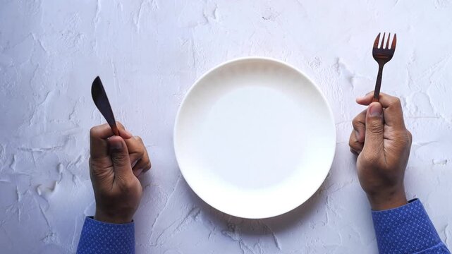 Top view of person holding knife and fork waiting for food at empty white plate on marble background
