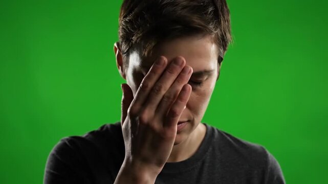 A young man covering his face with his hand in distress against a plain green background