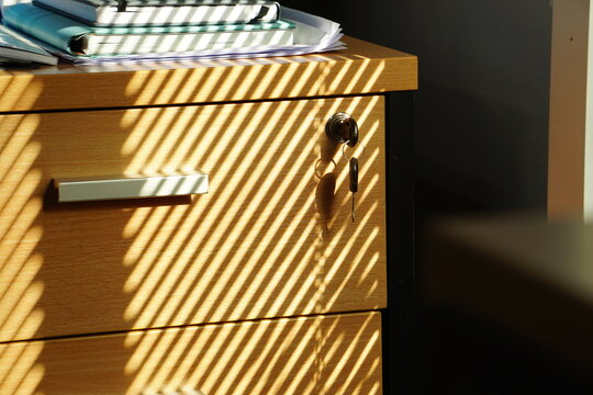 Natural light streams through window blinds, casting a rhythmic pattern of shadows across a wooden cabinet with a key inserted in the lock.
