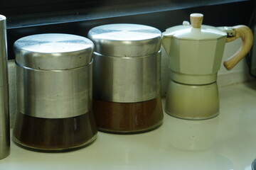 A classic Moka Pot stands next to two airtight canisters filled with dark coffee beans.