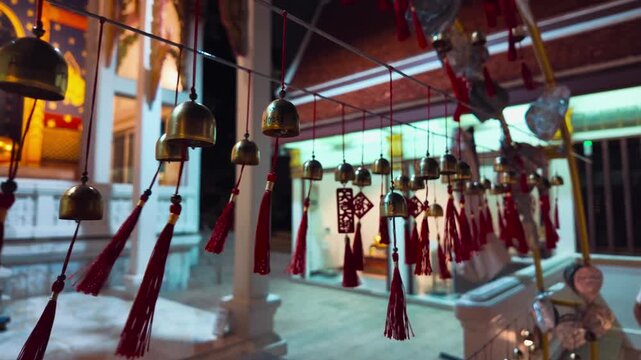 Row of bronze wind bells hanging in Thai Buddhist temple garden, Traditional sacred temple bells along railing with tropical foliage 4k