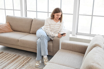 Obraz premium Young woman using mobile phone on grey couch near window at home