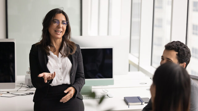 Mature 60s businesslady conducting informal meeting or briefing with team members, smiling, exchanging work-related ideas or thoughts, making decisions together, engaged in teamwork in coworking space