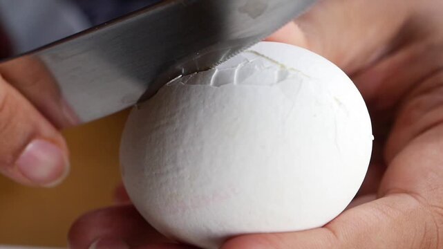Close up video of hands holding a white egg and carefully peeling its shell with a knife, showcasing the process of preparing a hard boiled egg