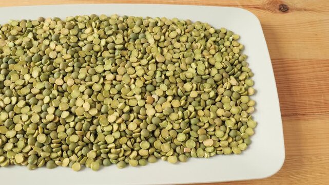 Dry green split peas slowly moving on a white plate over a wooden backgroundю