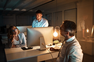 Fototapeta premium Computer, night and talking with business people in office together for deadline or overtime. Conversation, planning and working late with employee group in dark workplace for project management