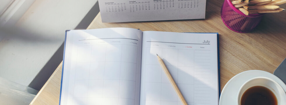 Agenda, planner book, calendar, pencil, laptop, and cup of coffee placed on business office desk. Diary for organizer to plan timetable, daily appointment, and management job in room-banner image.  