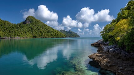 Obraz premium Serene Tropical Landscape with Lush Mountains, Clear Water, Bright Clouds, and Rocky Shoreline Under a Blue Sky