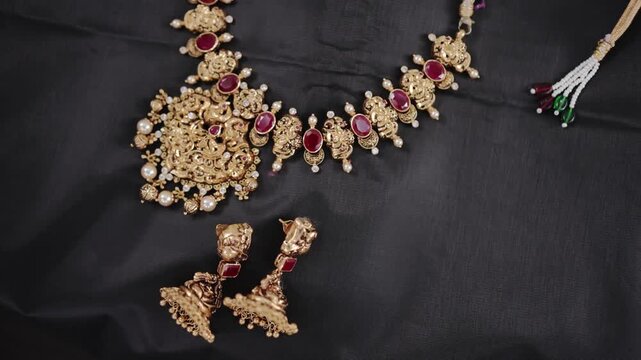 Top-down view of a luxurious traditional Indian bridal gold temple jewelry set featuring a ruby necklace and jhumka earrings on a dark textured fabric.