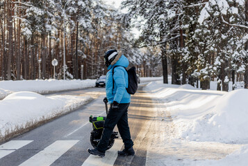 Person walking near electric unicycle monowheel crossing snowy winter street crosswalk in forest area wearing blue jacket helmet backpack showing alternative eco-friendly urban commute mobility © MarijaBazarova