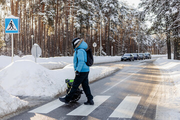 Person walking near electric unicycle monowheel crossing snowy winter street crosswalk in forest area wearing blue jacket helmet backpack showing alternative eco-friendly urban commute mobility © MarijaBazarova
