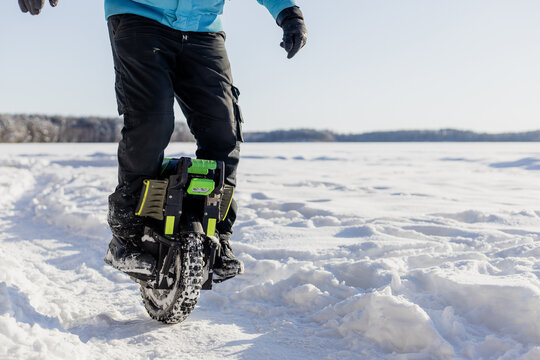 Person riding electric unicycle monowheel on frozen snow-covered lake field in winter landscape with blue sky forest background, extreme outdoor adventure sport activity